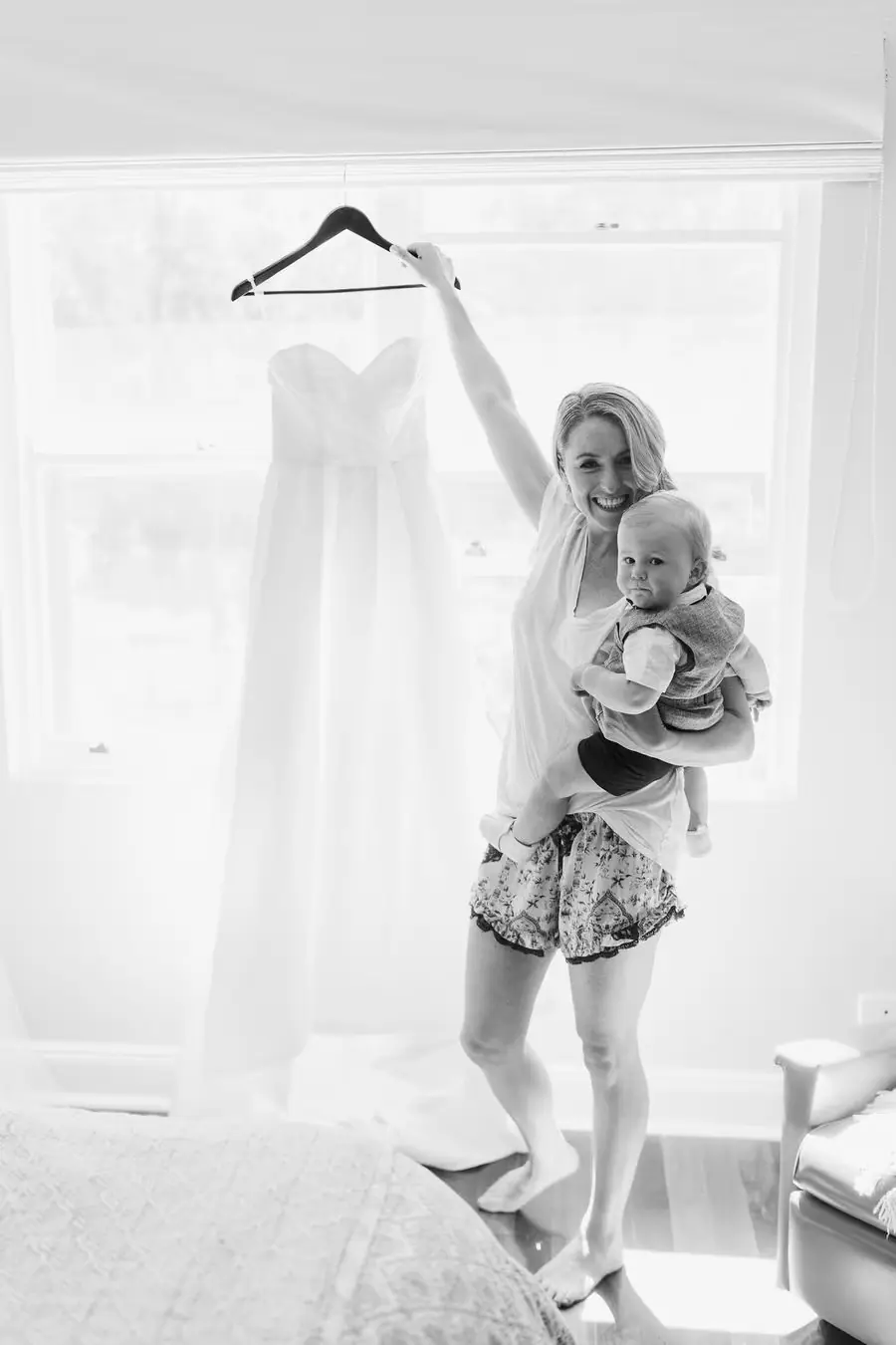 Black and white photograph of a woman holding a baby and a hanging wedding dress.