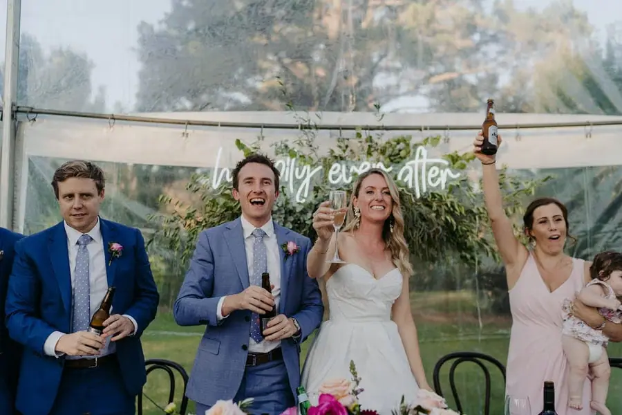 Couple and guests raise drinks for a toast beneath a reception sign.