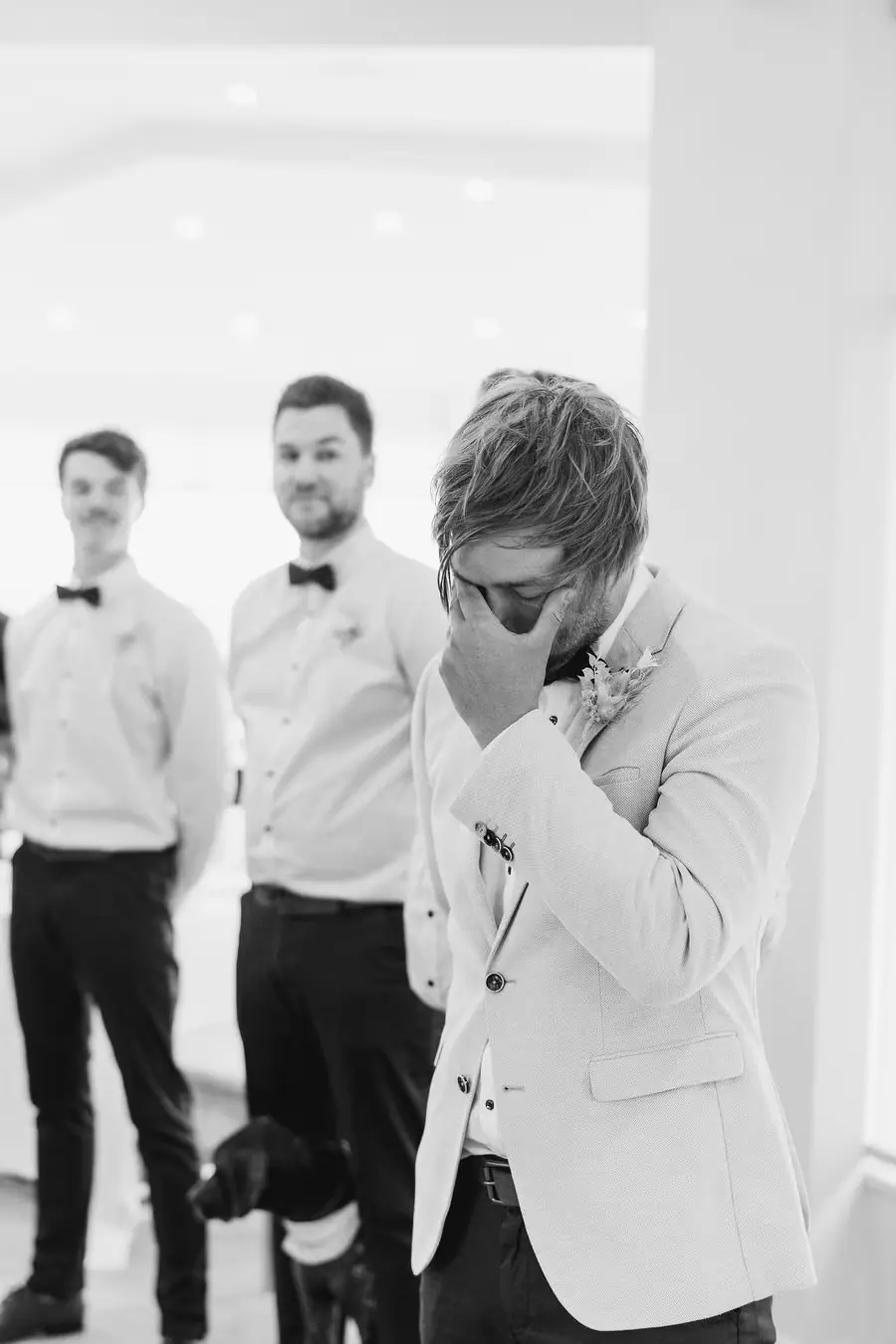 Black and white photograph of a man wiping his eye with two groomsmen behind him.