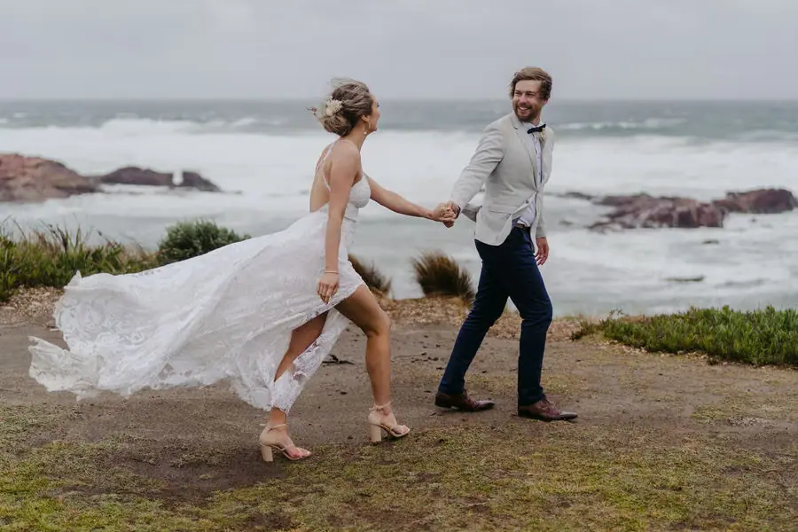 Couple walks hand in hand along a rocky coastline with the bride's train blowing behind her.