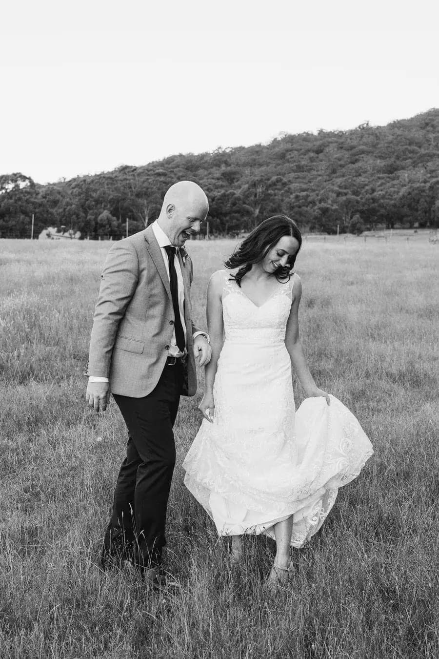 Black and white photograph of the couple walking through grass with the bride holding her skirt.
