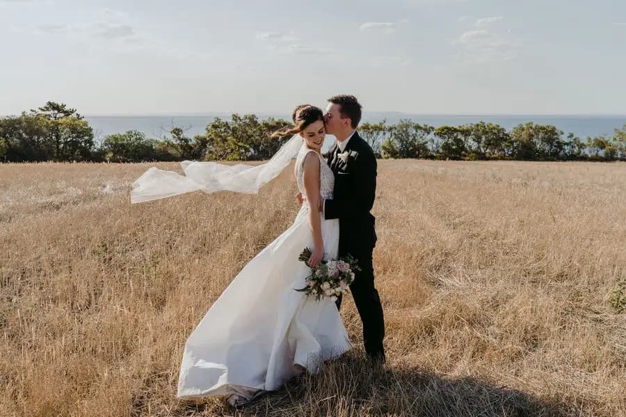 Couple embraces in a dry coastal field with the bride's veil blowing sideways.