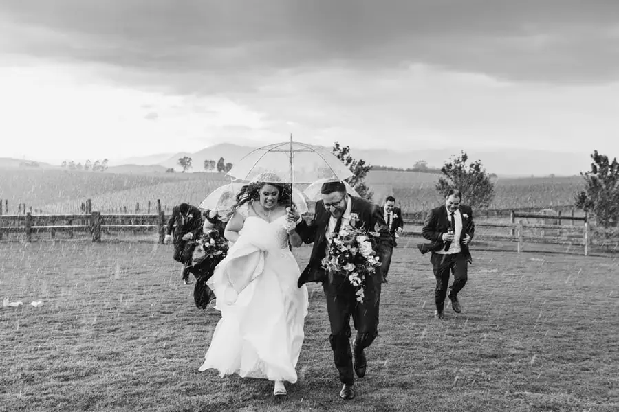 Black and white photograph of the couple and wedding party running beneath clear umbrellas.