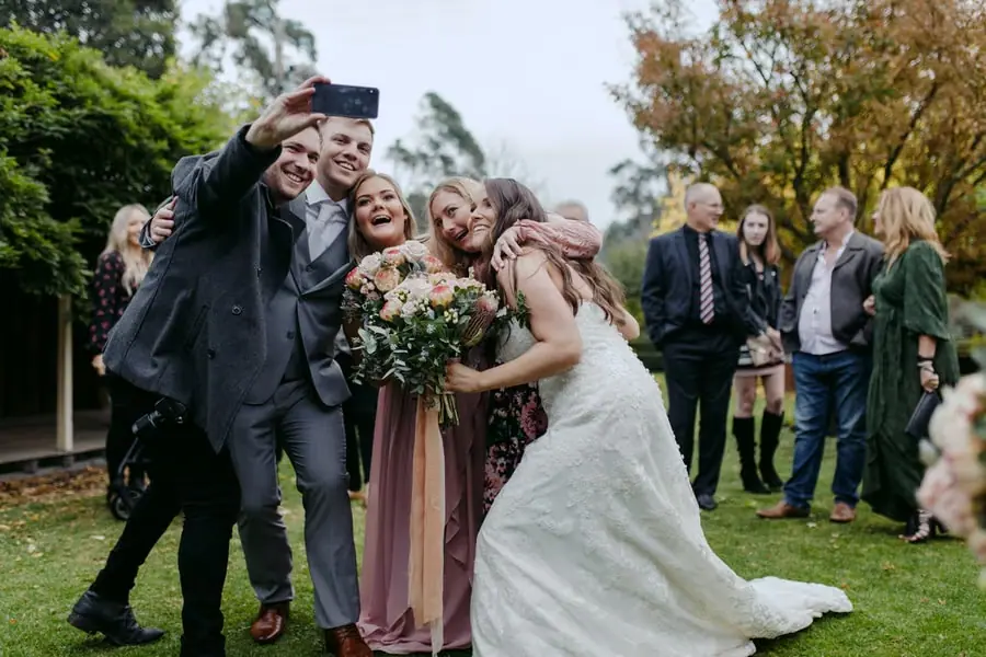 Bride and guests lean together for a phone selfie outdoors.
