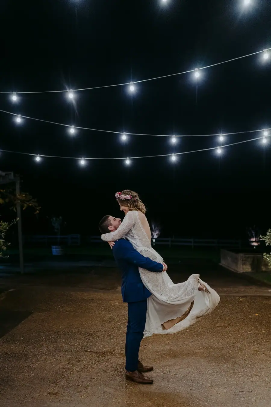 Groom lifts the bride beneath string lights at night.