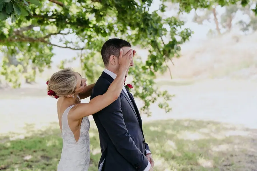 Bride reaches up to the groom during their first look beneath a leafy tree.
