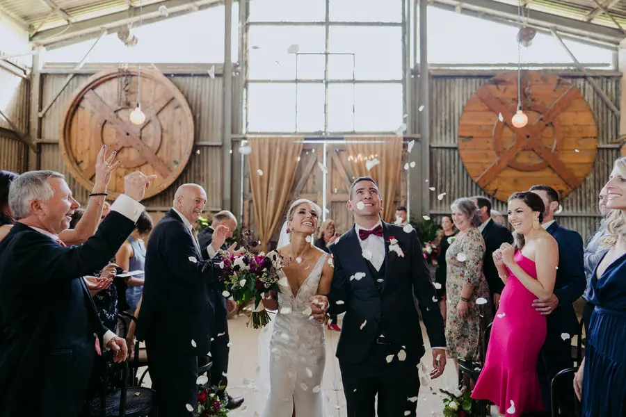 Couple celebrates amid confetti inside a reception venue with large wooden reels on the wall.