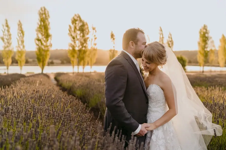 Groom kisses the bride's forehead beside rows of flowering plants at sunset.