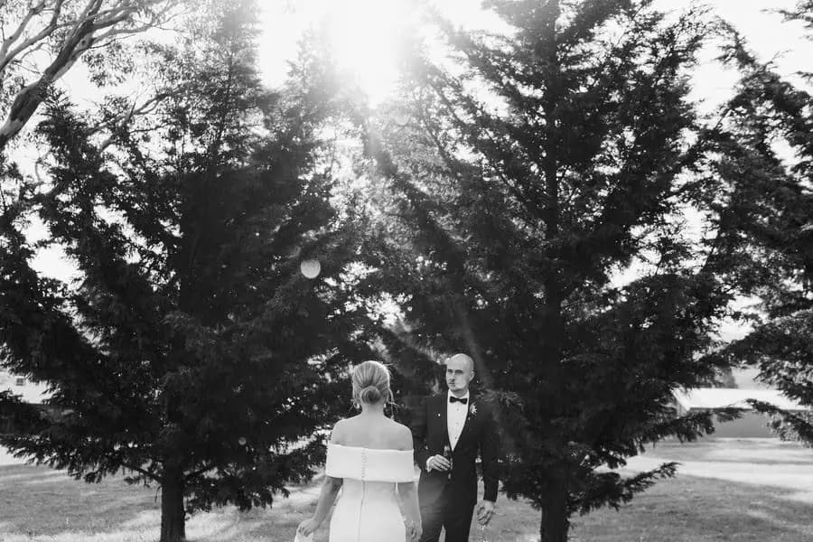 Black and white photograph of the bride approaching the groom beneath tall trees.