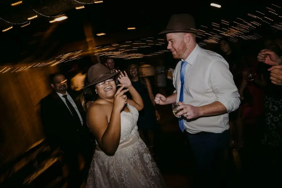 Couple dances wearing brimmed party hats among reception guests.