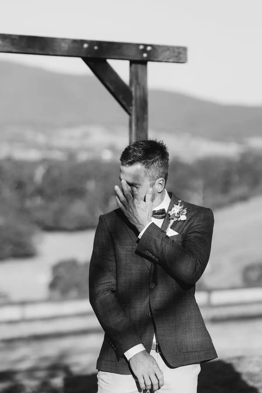 Black and white photograph of the groom wiping his eye beside a wooden ceremony arch.