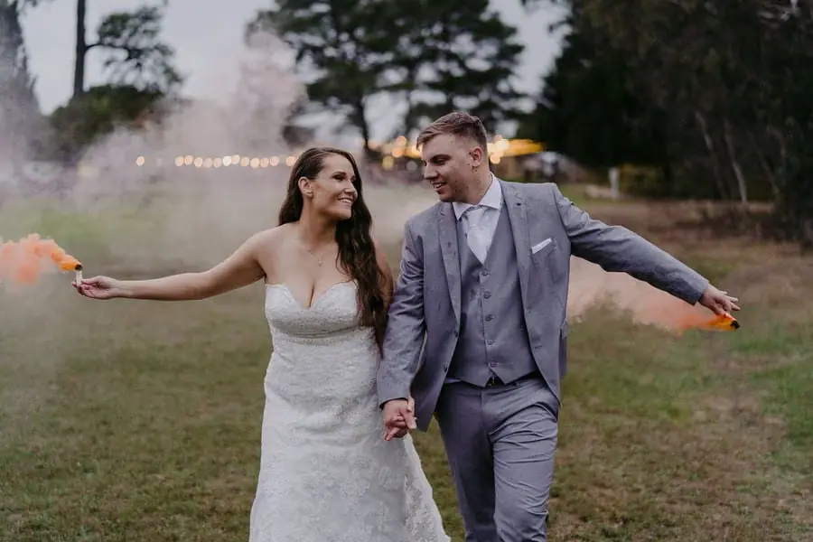 Couple holds hands while coloured smoke drifts from objects in their outstretched hands.