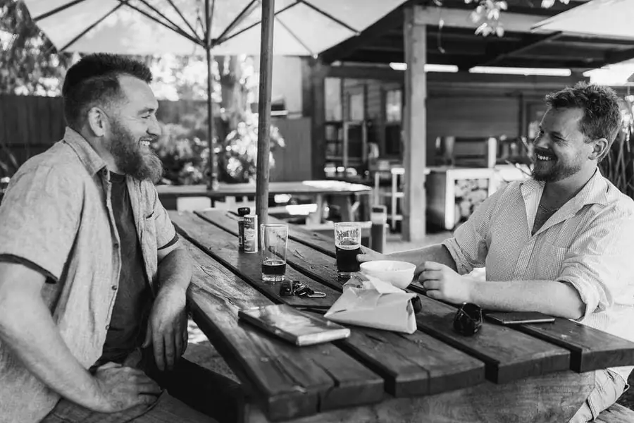 Black and white photograph of two men laughing over drinks at a wooden table.