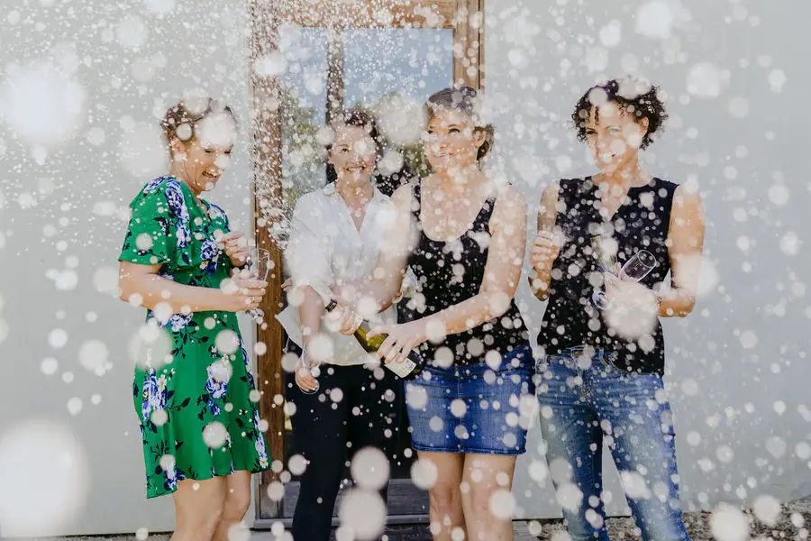 Women laugh as champagne sprays toward the camera indoors.