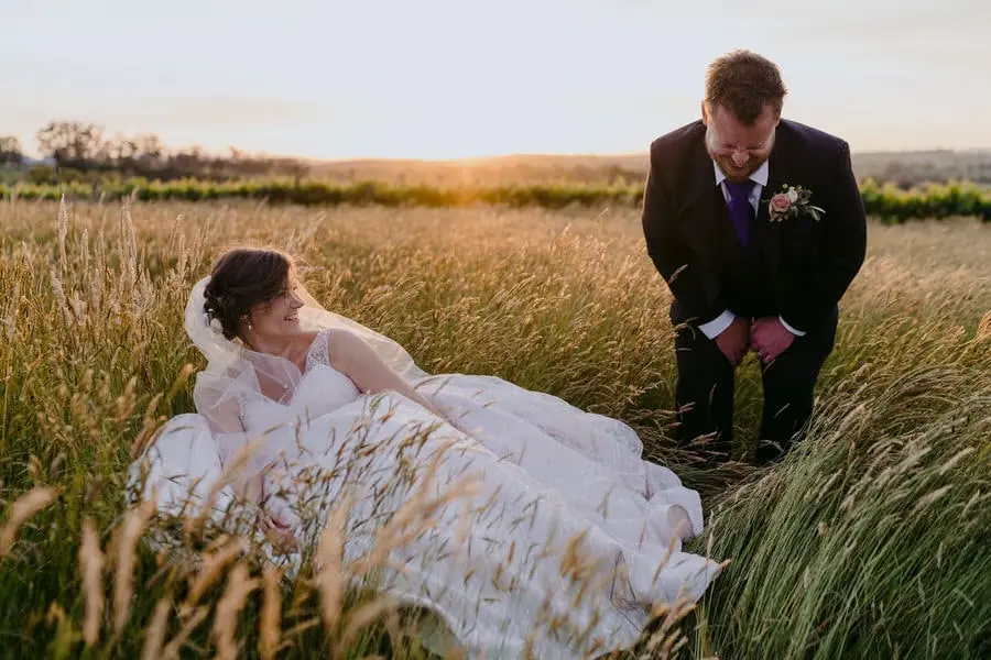 Bride sits in long grass laughing toward the groom standing beside her.