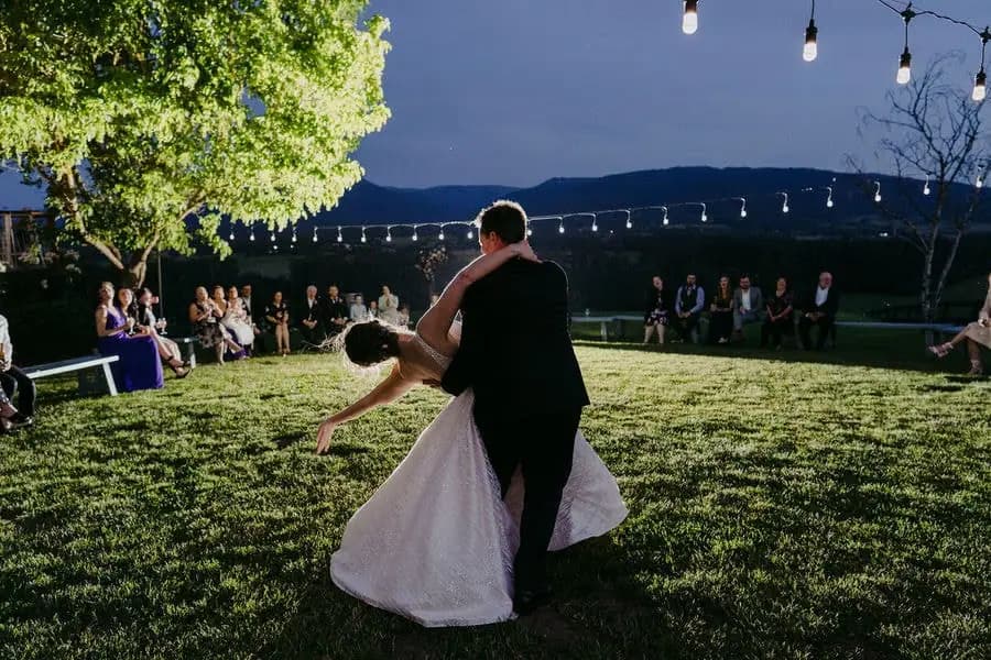 Groom dips the bride on a lawn beneath evening string lights.