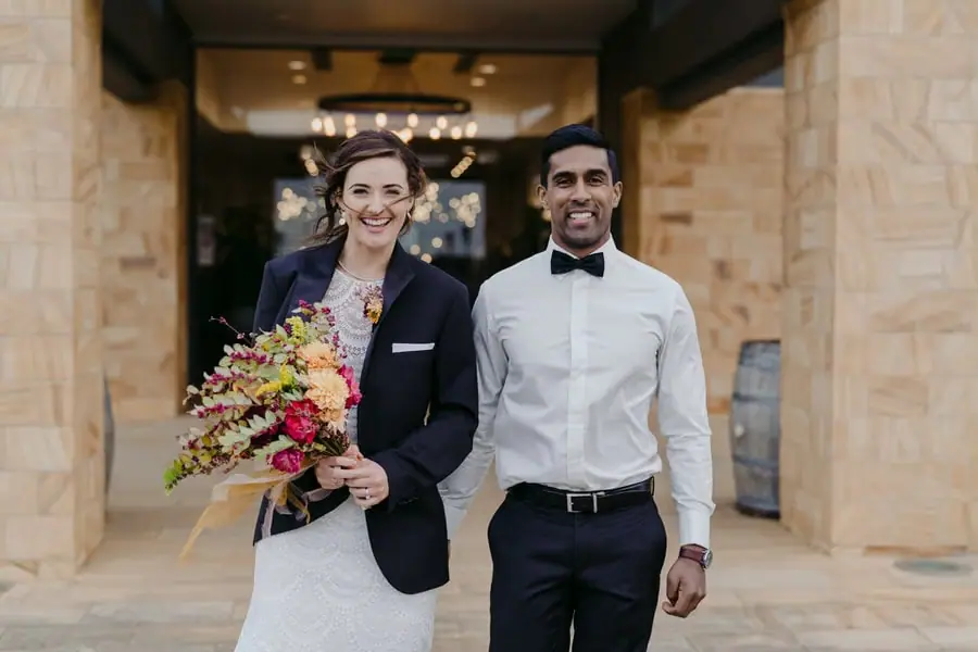 Couple smiles outside a stone building with the bride wearing a dark jacket.