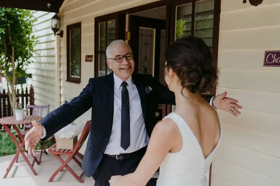 Older man opens his arms to greet the bride beside a house.