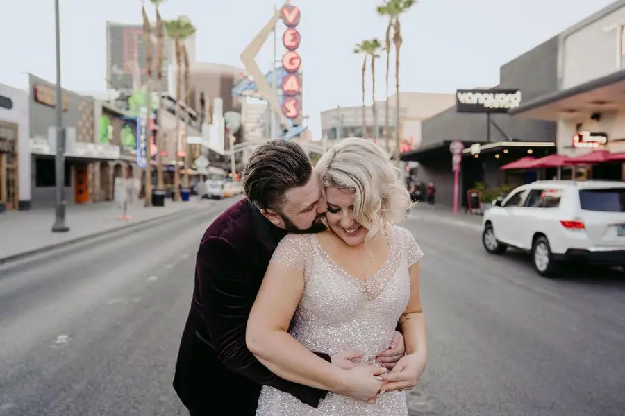 Groom embraces and kisses the bride from behind on a city street.