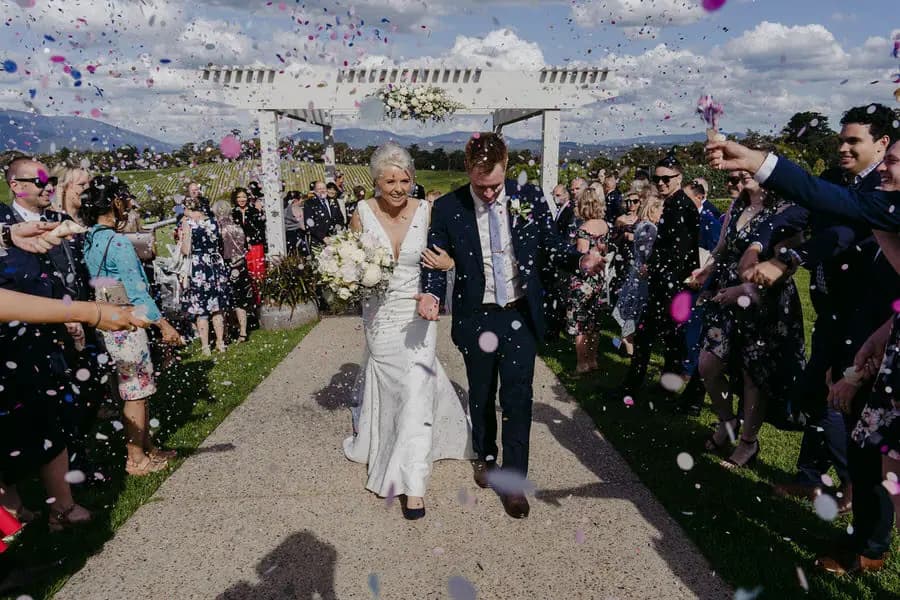 Newlyweds walk through a shower of confetti along an outdoor aisle.