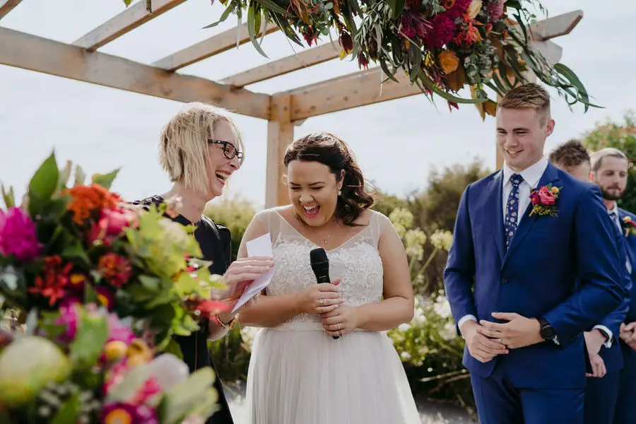 Bride laughs while holding a microphone beside the groom beneath a floral arch.