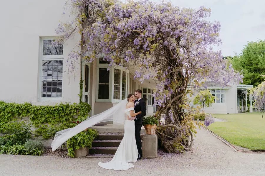 Couple embraces outside a house covered in purple flowering vines.