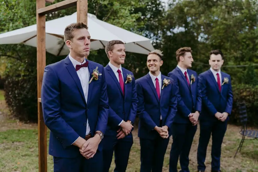 Groom and four groomsmen wait beside the outdoor ceremony arch.