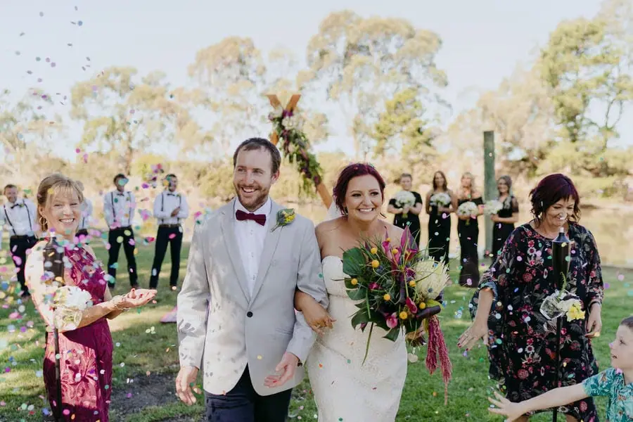 Couple walks through a shower of confetti with guests beside them.