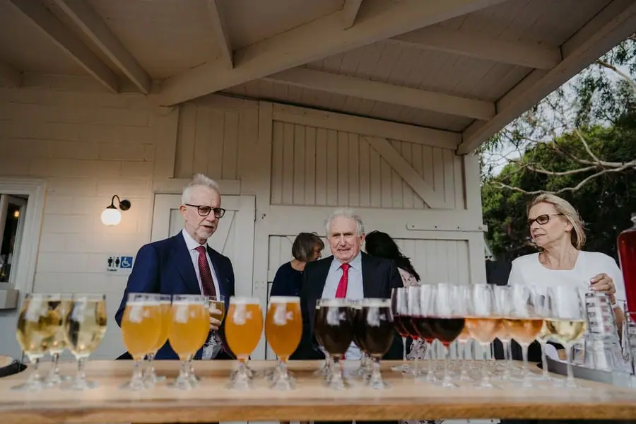Guests stand behind rows of filled wine glasses on a serving table.