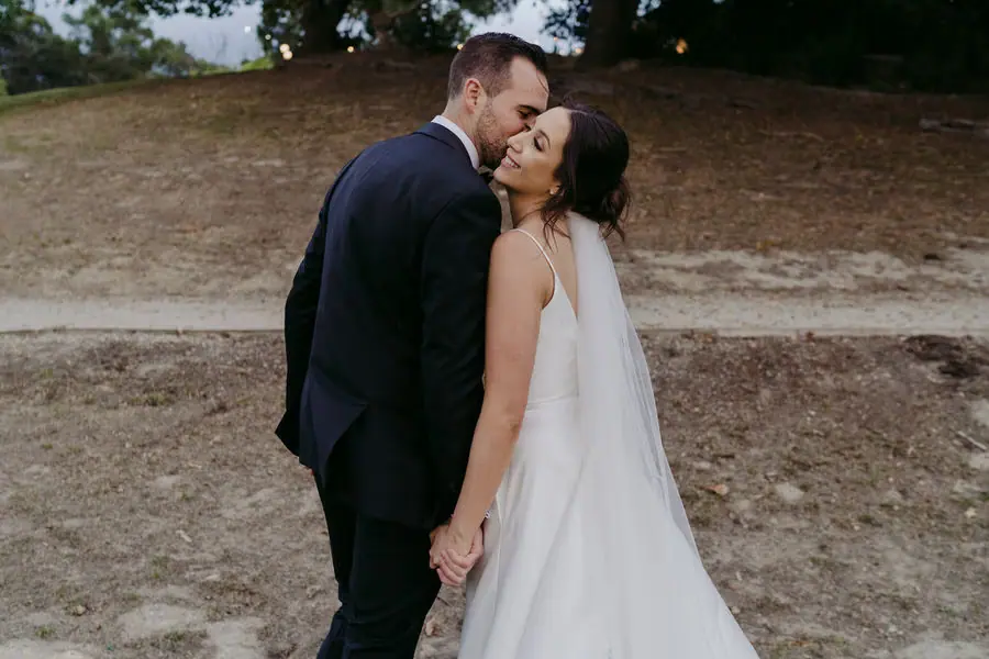 Couple kisses while holding hands beside a wooded slope.