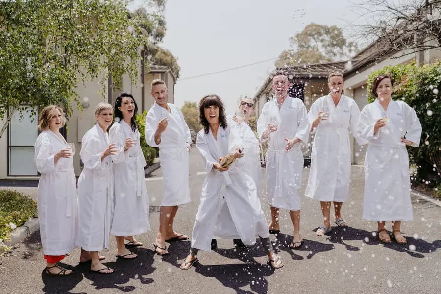 Women in white robes laugh as champagne sprays outdoors.