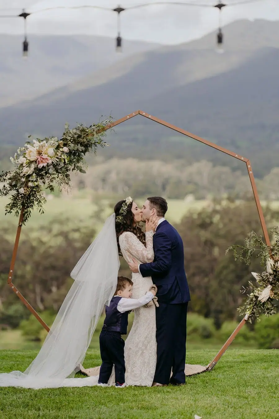 Couple kisses beneath a geometric floral arch overlooking hills.