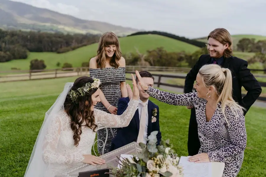 Guests reach toward the seated bride during outdoor wedding paperwork.