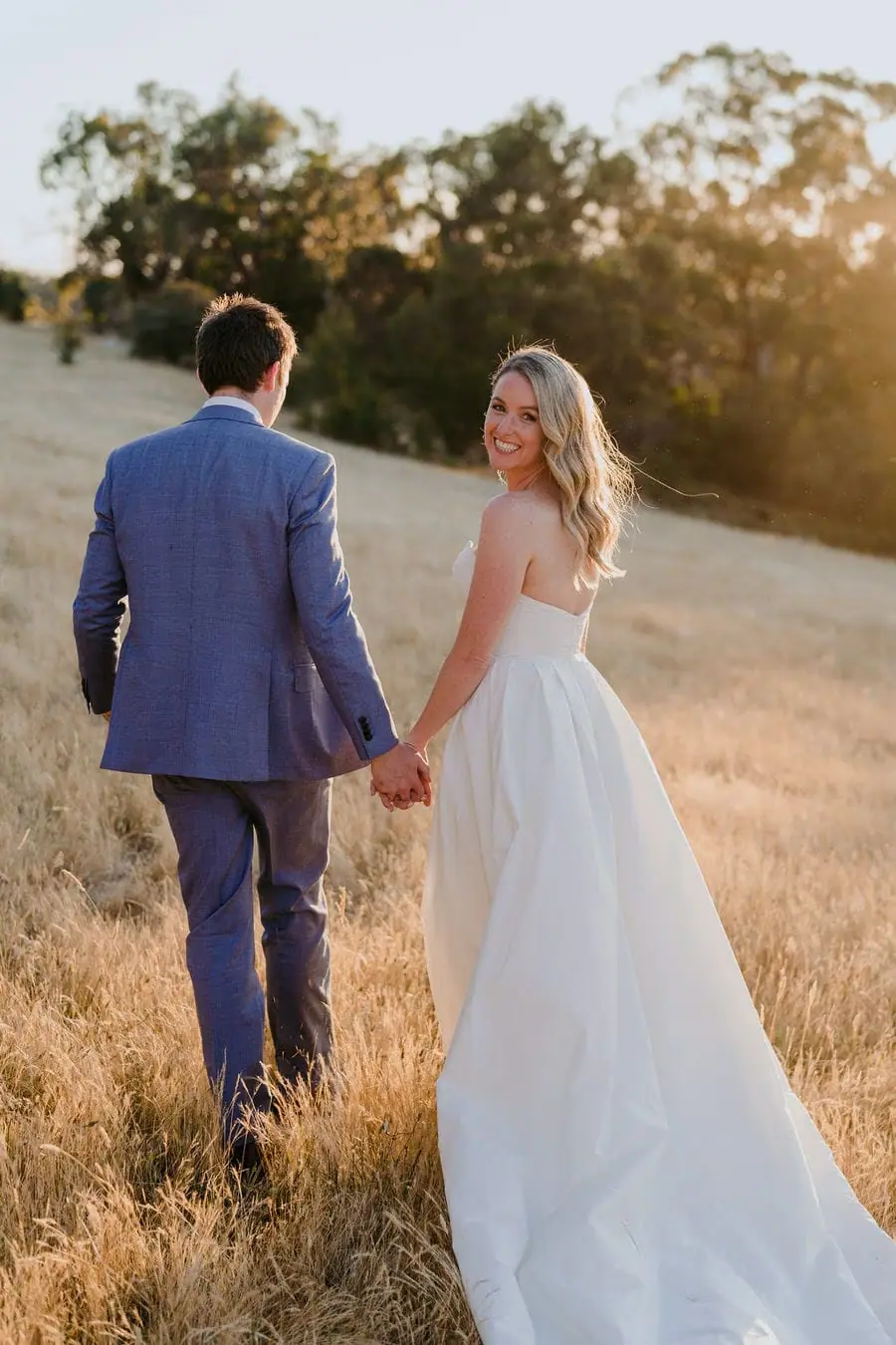 Rear view of the couple holding hands in a sunlit dry field as the bride looks back.