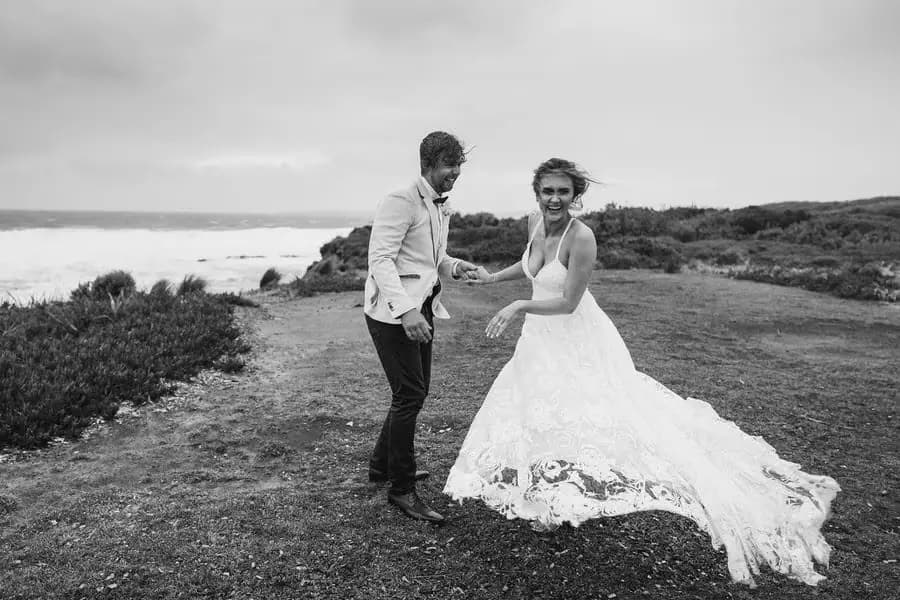 Black and white photograph of the couple standing beside the coastline with the bride's lace train spread out.