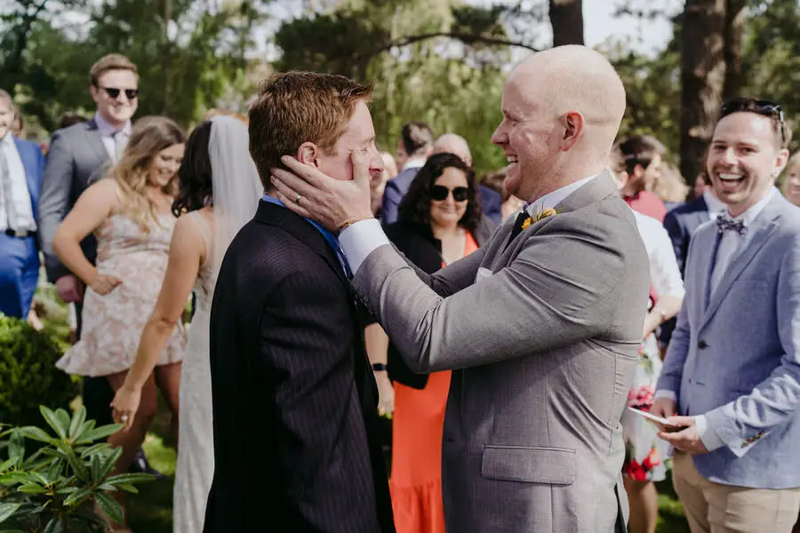 Older man holds the groom's face in his hands as guests smile nearby.