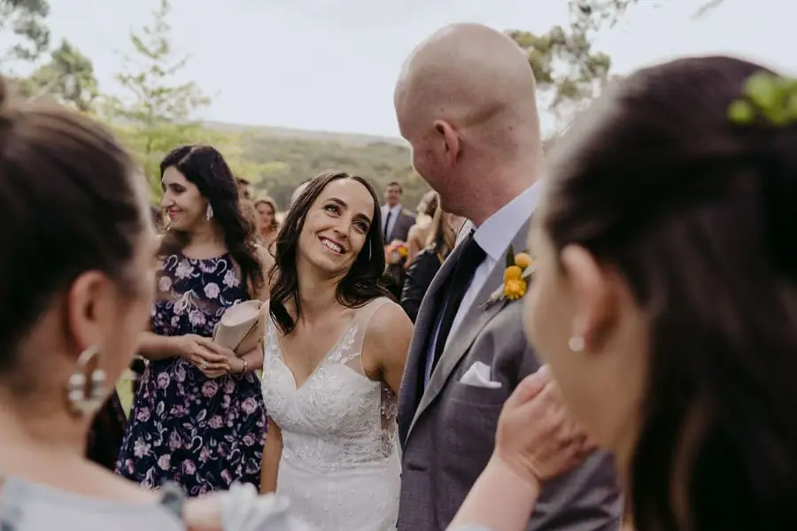 Bride smiles toward a suited guest among wedding guests outdoors.