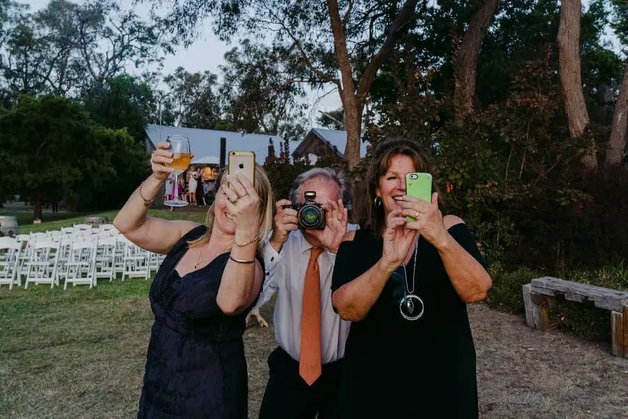Guests hold up phones and a camera for photographs outdoors.