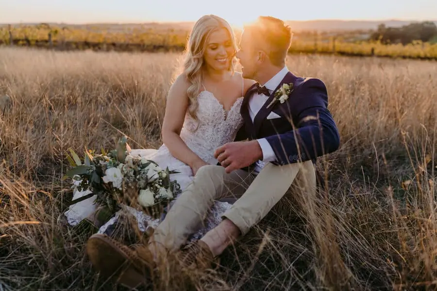 Couple sits in long grass at sunset with the bouquet beside them.