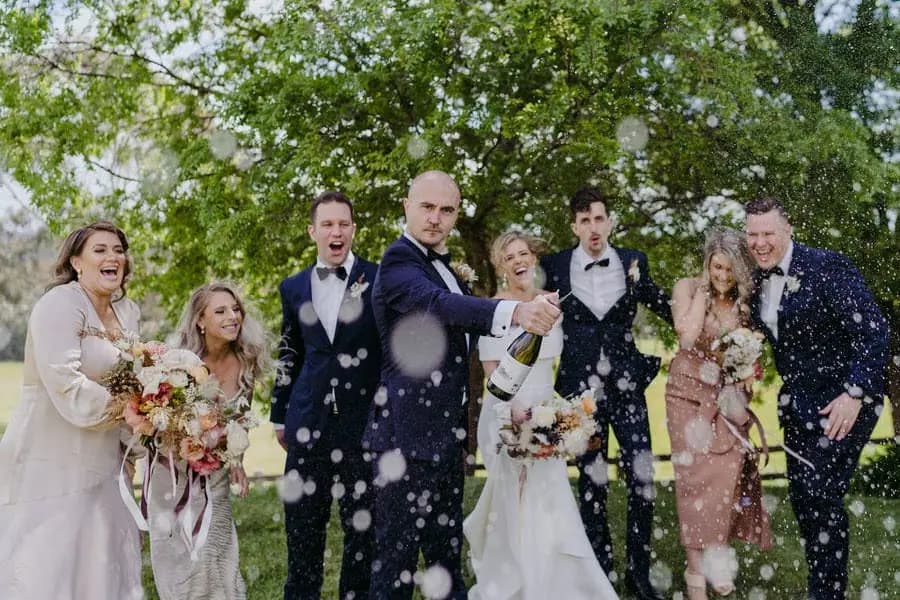 Wedding party celebrates as champagne sprays toward the camera beneath a leafy tree.