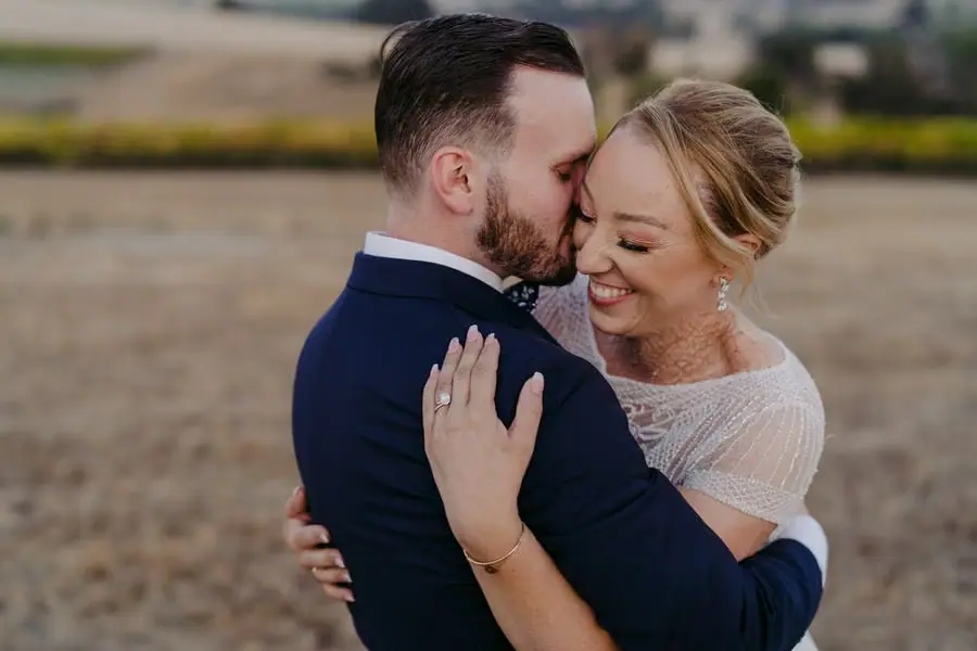 Groom kisses the smiling bride's forehead in a dry field.