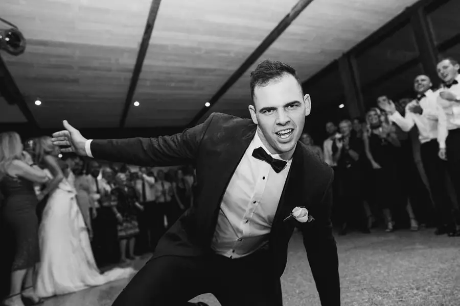 Black and white photograph of the groom dancing toward the camera with guests behind him.