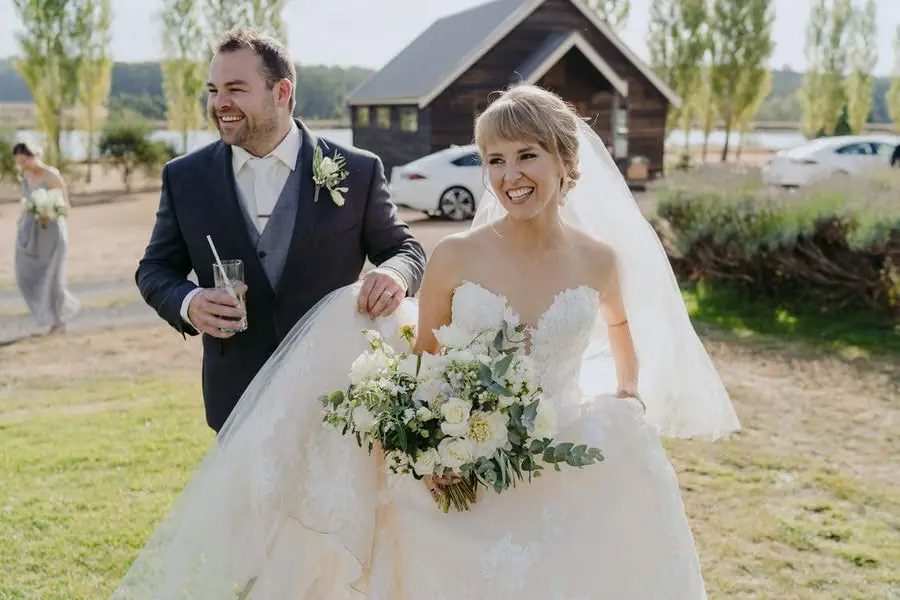 Couple walks together across a sunny lawn with the groom holding a drink.