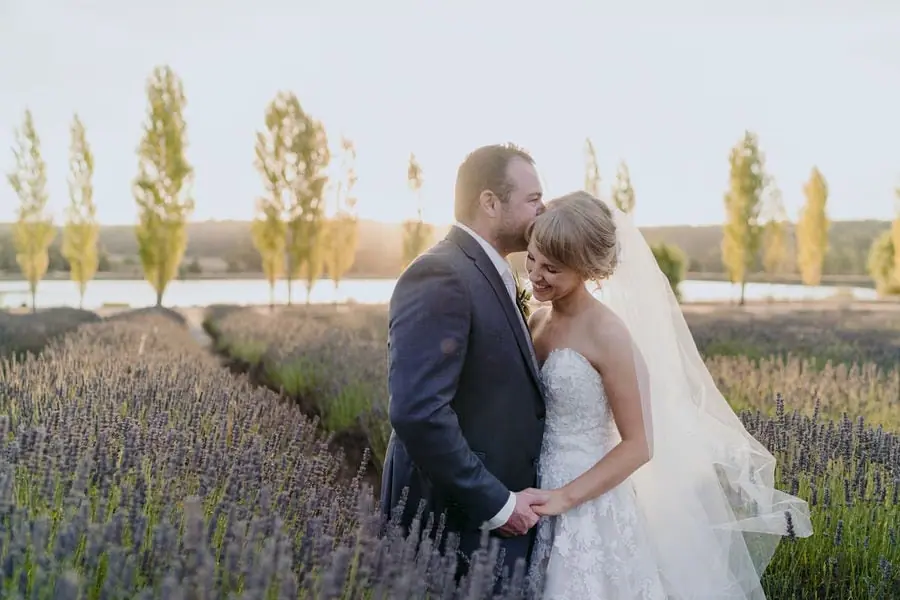 Couple holds hands among flowering plants with sunset light behind them.