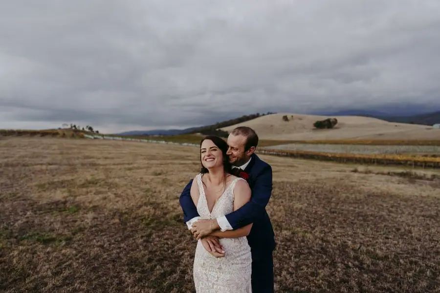 Groom embraces the smiling bride from behind beneath a dark cloudy sky.