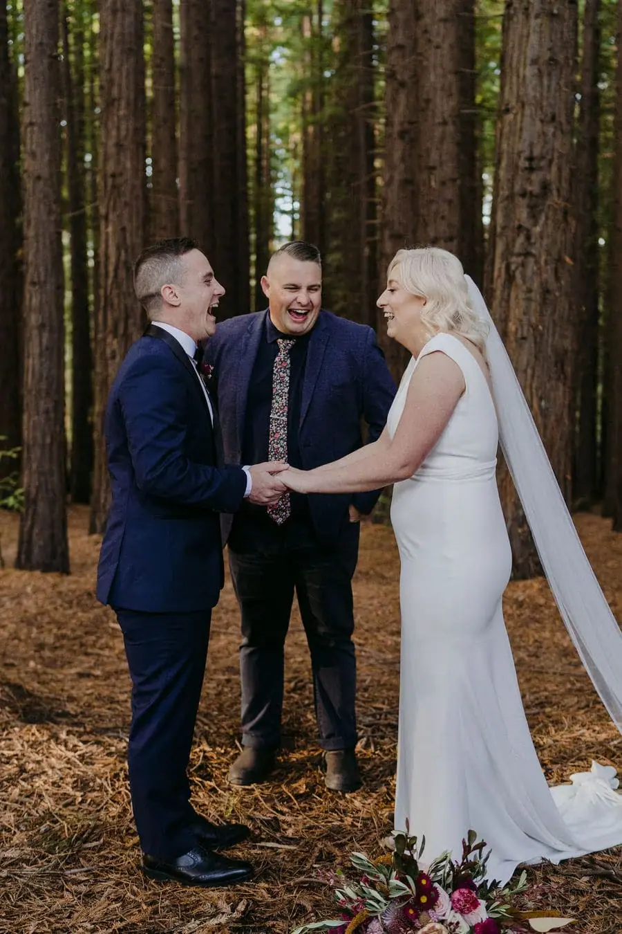 Couple holds hands beside a celebrant among tall forest trees.