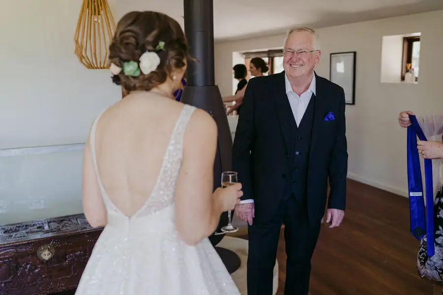 Older man smiles toward the bride while holding a drink indoors.