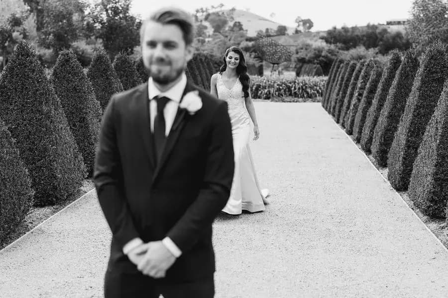 Black and white photograph of the groom waiting as the bride approaches along a hedged path.