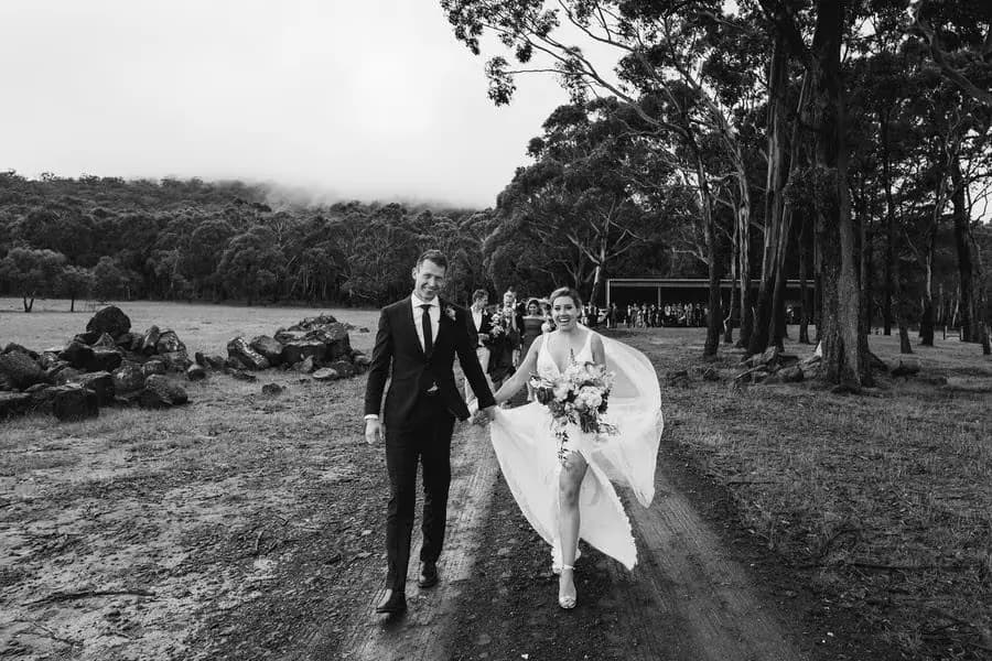 Black and white photograph of the couple walking through a field with the bride gathering her skirt.