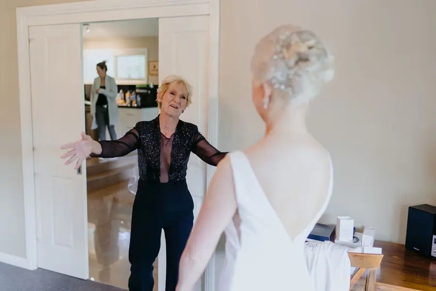 Older woman opens her arms to greet the bride indoors.